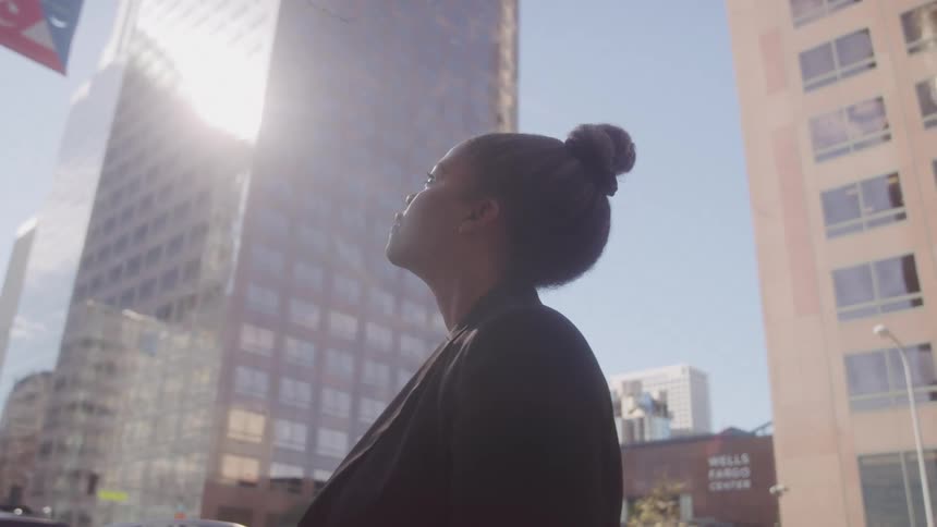A woman stands in front of tall buildings as sunlight reflects off the glass faade