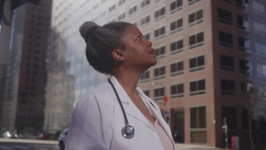 A doctor with a stethoscope around her neck stands in a city street gazing up at the surrounding tall buildings