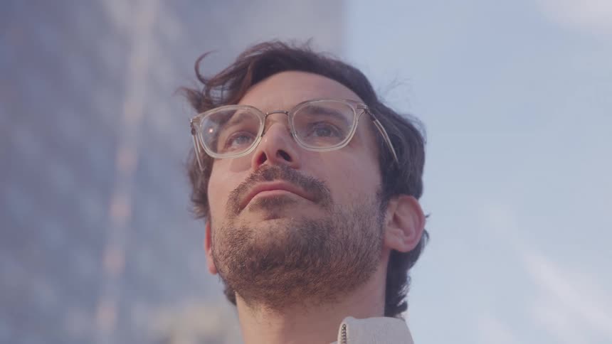 A young bearded man wearing glasses is standing in the middle of the city surrounded by highrise buildings at midday