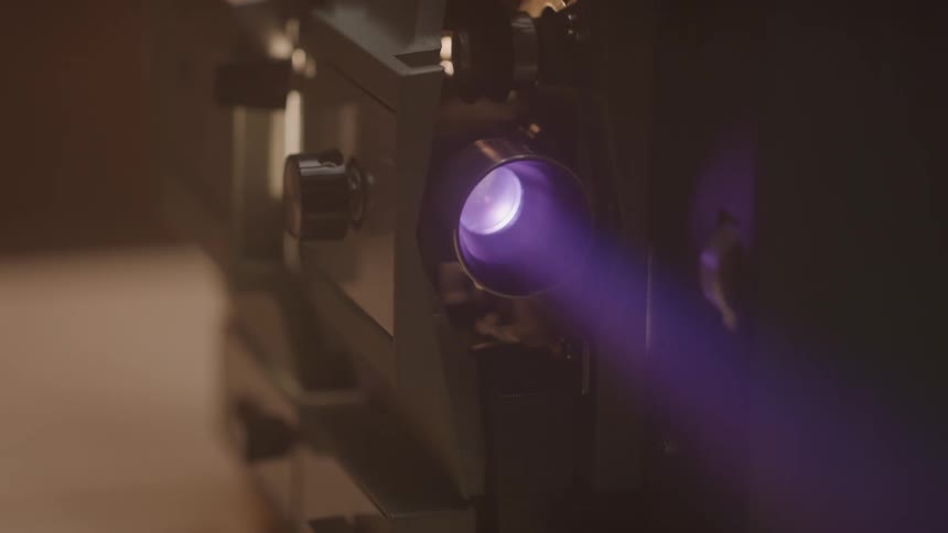 A closeup view focuses on the lens of an old film projector emitting a powerful purplehued light beam into a dark room the light pulses gently suggesting the mechanical whirring of the projector as it casts images onto a screen beyond the frame