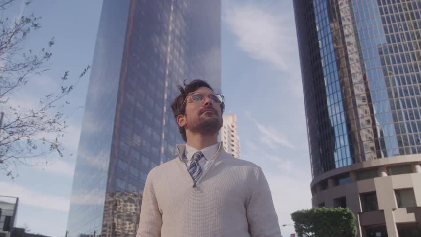 A man wearing glasses a light sweater and a tie stands outdoors gazing upwards with a contemplative expression he is framed between two towering modern buildings with reflective facades suggesting an urban environment