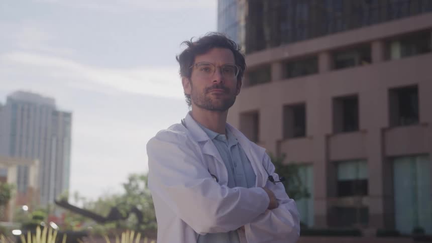 A male doctor with tousled dark hair and glasses stands outdoors with his arms crossed wearing a white lab coat over a light blue shirt he looks directly at the camera with a serious yet approachable expression against a blurred backdrop