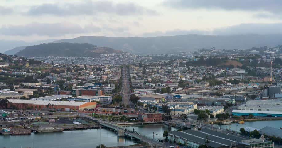 A sprawling urban landscape features a long street cutting through buildings and residential areas with a bridge crossing a narrow channel in the foreground and hills in the distance