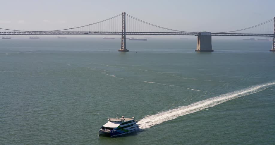 A highangle view captures a modern ferry boat creating a white wake as it speeds across a vast body of water in the background a large suspension bridge spans the water with other ships dotted across the horizon under a bright sky
