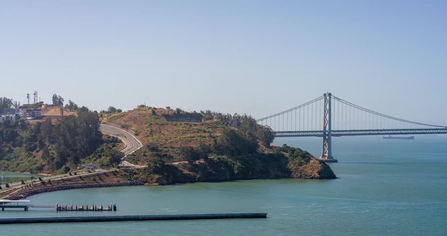 A winding road leads across a hilly island to a red suspension bridge spanning the calm blue waters of the bay