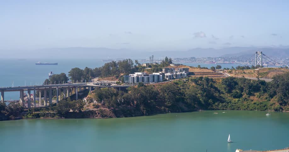 A long curving bridge connects to a lush island with buildings overlooking a calm bay with a sailboat and a distant city shoreline