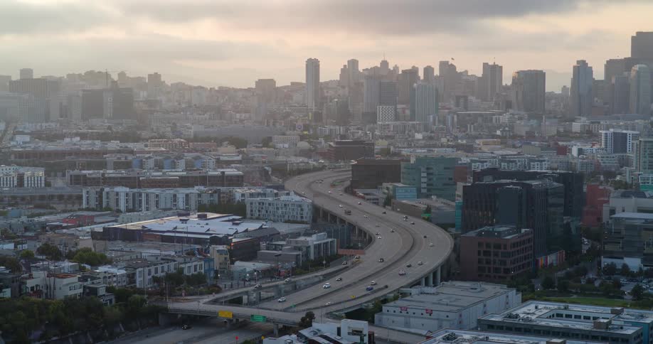 A city skyline view at midday features an overhead bridge spanning through the city as cars pass by