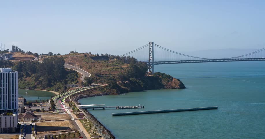 A curving coastal road leading to a bridge with a small pier jutting into the calm bay below and a tall building