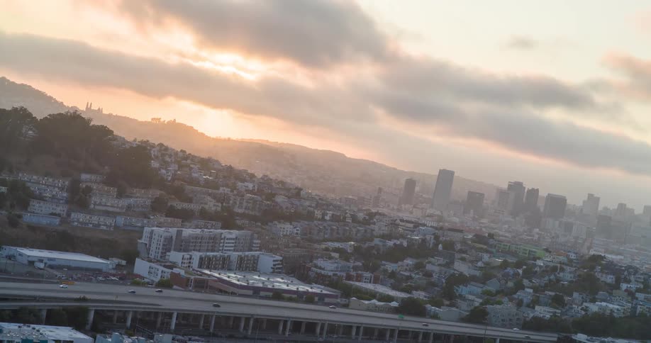 Light traffic travels on a highway passing through a residential area while the rising sun shines over the foggy city