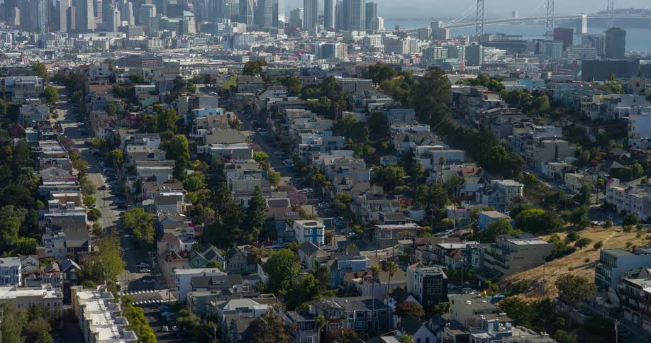 An aerial shot reveals a dense residential neighborhood with numerous houses and green trees lining the streets stretching up a hillside in the distance a prominent city skyline with skyscrapers and a large suspension bridge can be seen under a bright