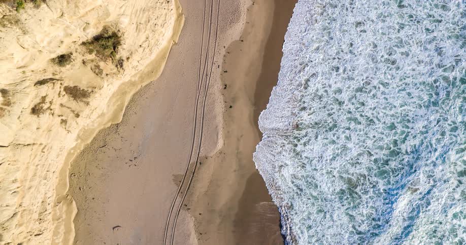 Tire tracks curve across a wide sandy beach toward the shoreline where waves break next to a steep cliff