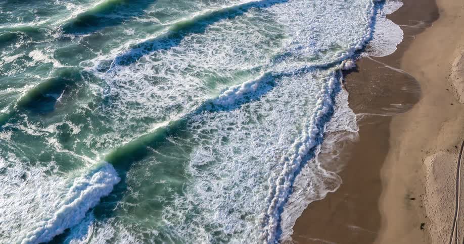 Waves crash onto a sandy beach creating intricate patterns of foam and water