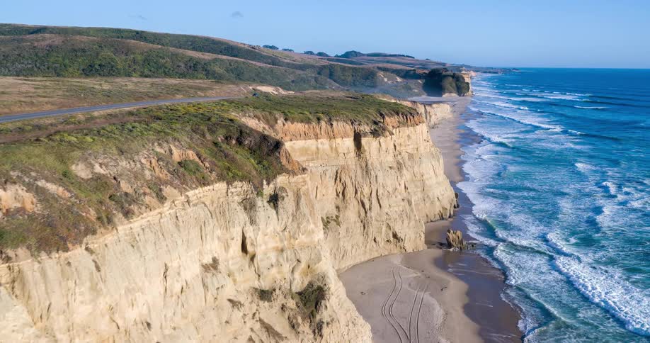 A car drives along a rugged coastal road where steep lightbrown cliffs drop into a turquoise ocean with white waves crashing ashore