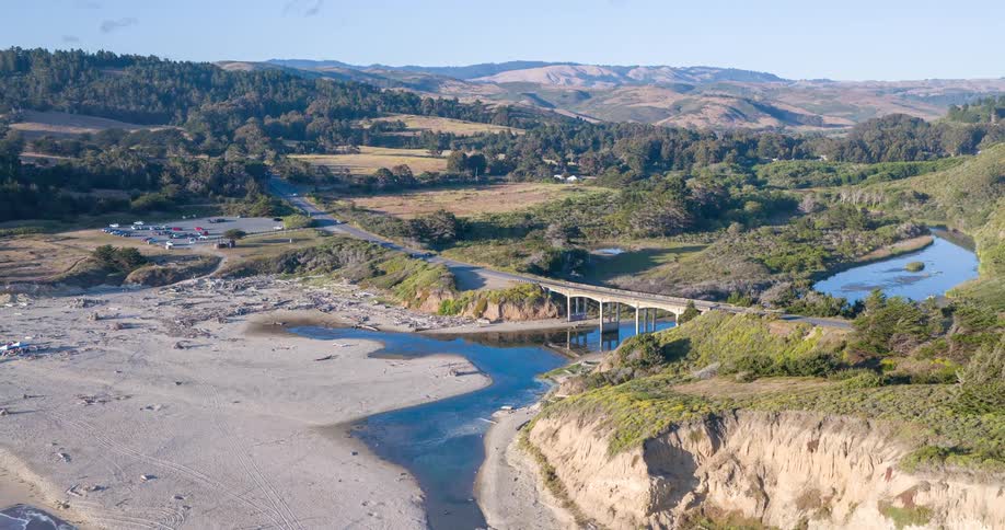 A scenic aerial view of a coastal landscape a long winding river or estuary flows through a valley eventually meeting a wide sandy beach a bridge spans the water with a road leading towards distant hills covered in lush green trees