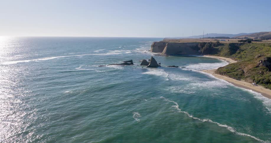 A coastal landscape shows light brown cliffs extending into the turquoise ocean with waves breaking on a sandy beach and a clear sky overhead