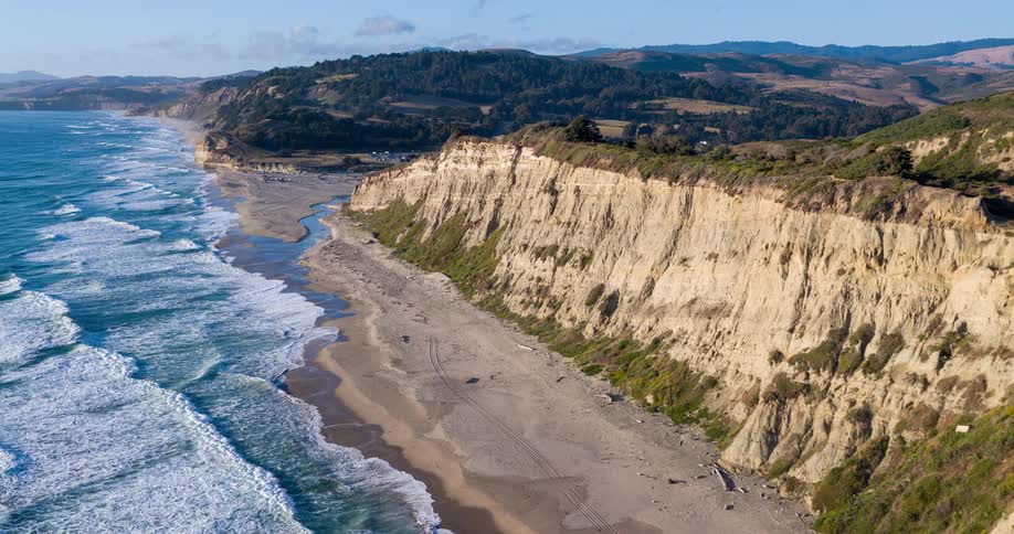 An ocean view from the shoreline shows a hill in the distance as waves hit the shore at midday