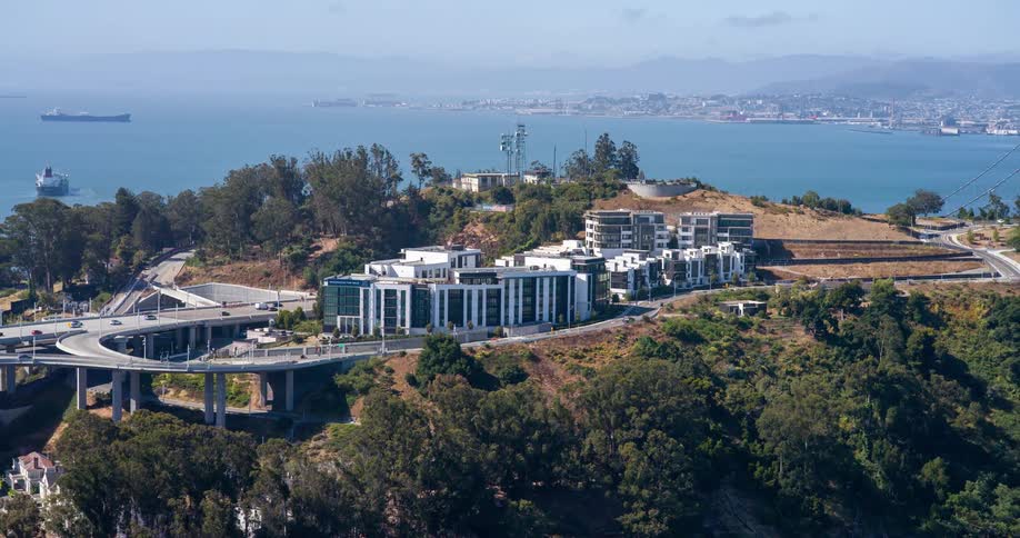 Cars driving past a collection of buildings atop a large hill