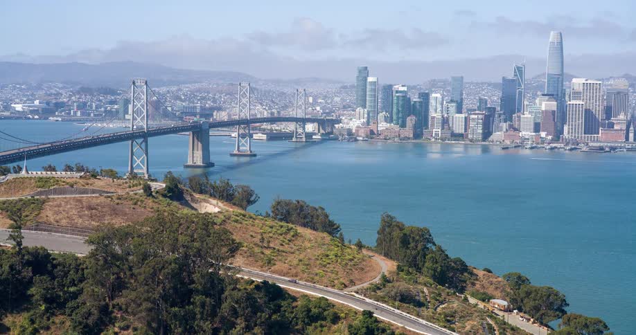 A bridge stretches over a river leading into a city filled with a series of buildings and skyscrapers at midday