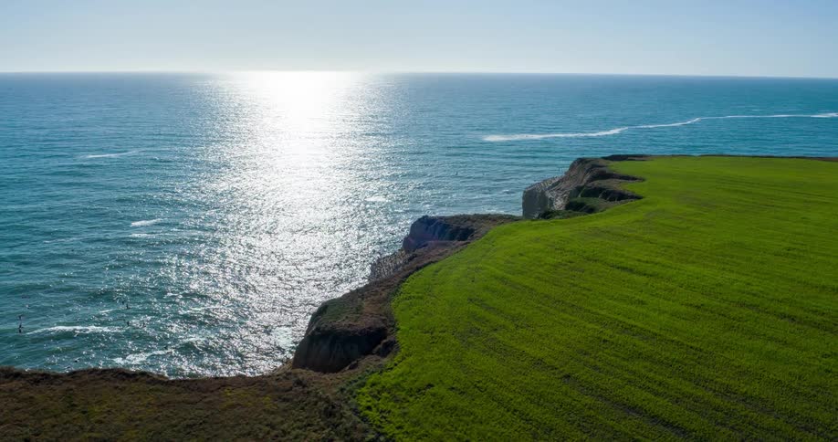 A rocky cliffside juts into a shimmering blue ocean beneath a clear sky with green grass along the top edge