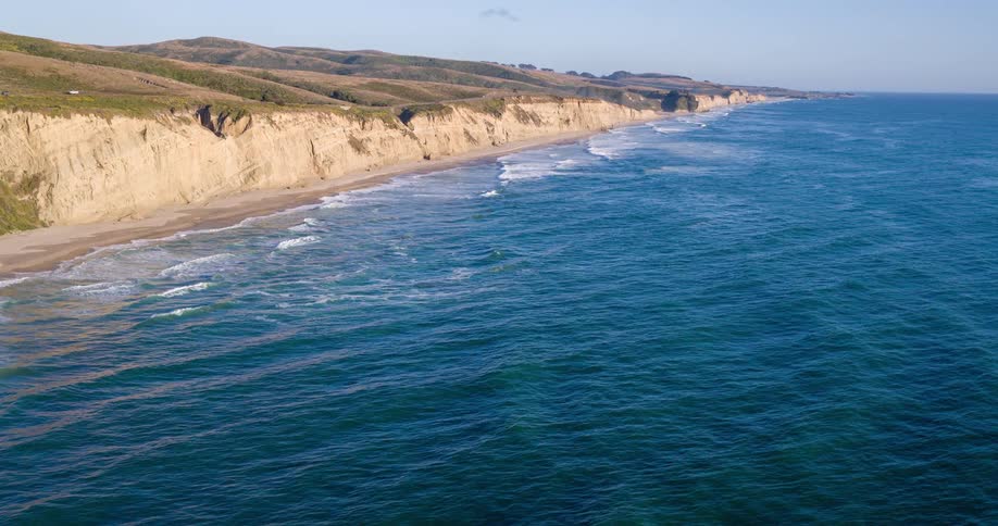 A breathtaking aerial view of a rugged coastline featuring a long stretch of sandy beach nestled against dramatic steep cliffs covered in sparse vegetation the deep blue ocean with its foamy white waves crashing onto the shore