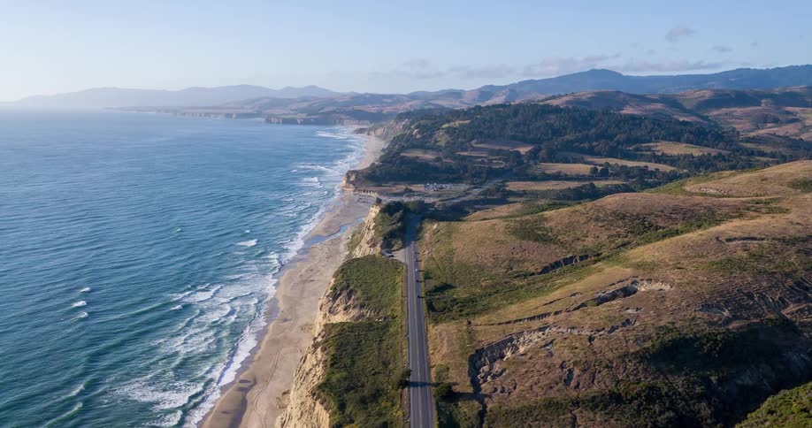 A stunning aerial view of a rugged coastline with a long straight road running parallel to the deep blue ocean waves crash onto the sandy beach at the base of towering greencapped cliffs with rolling hills visible in the distance under a clear sky