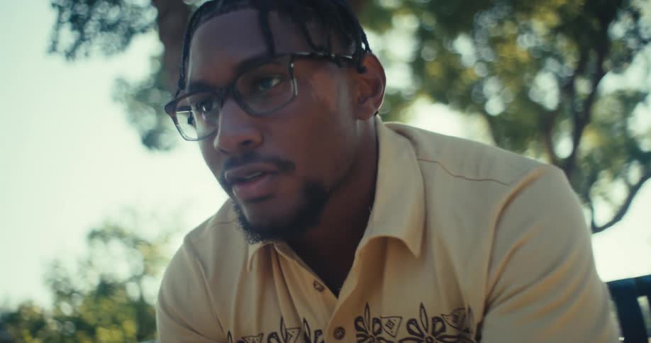 A black man talks on his phone while sitting in a sunlit park