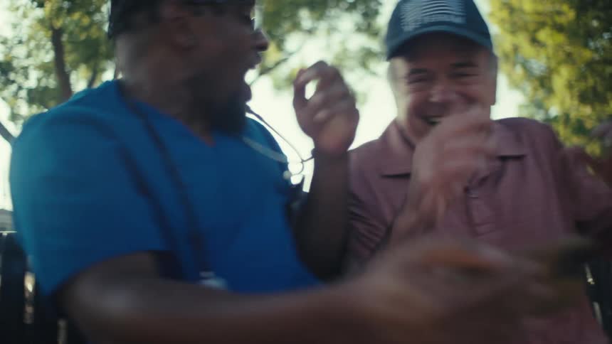 A doctor wearing a blue robe with a stethoscope over his shoulder is sitting on a bench in the park beside an elderly man both are filled with excitement as they watch a game on the mobile device held by the doctor at midday