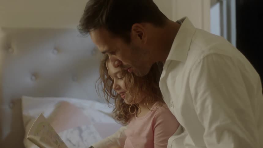 A young girl hops into bed as a father reads from a book