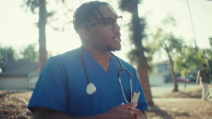 A healthcare worker in scrubs speaks with an older man in a sunlit residential area