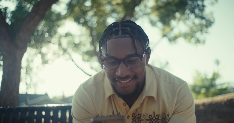 A young black man contemplates and smiles while texting on a phone outdoors during daytime