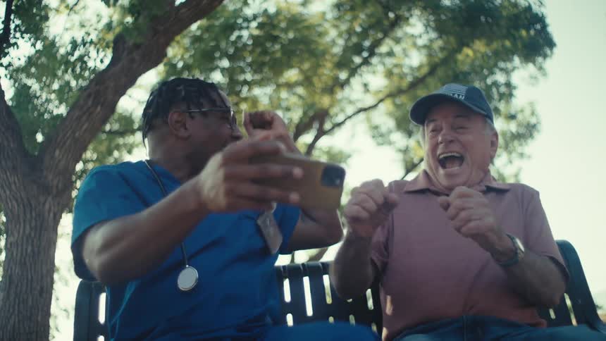 Male nurse and an elderly gentleman both sitting on a bench outdoors celebrating together happily watching something on a smartphone