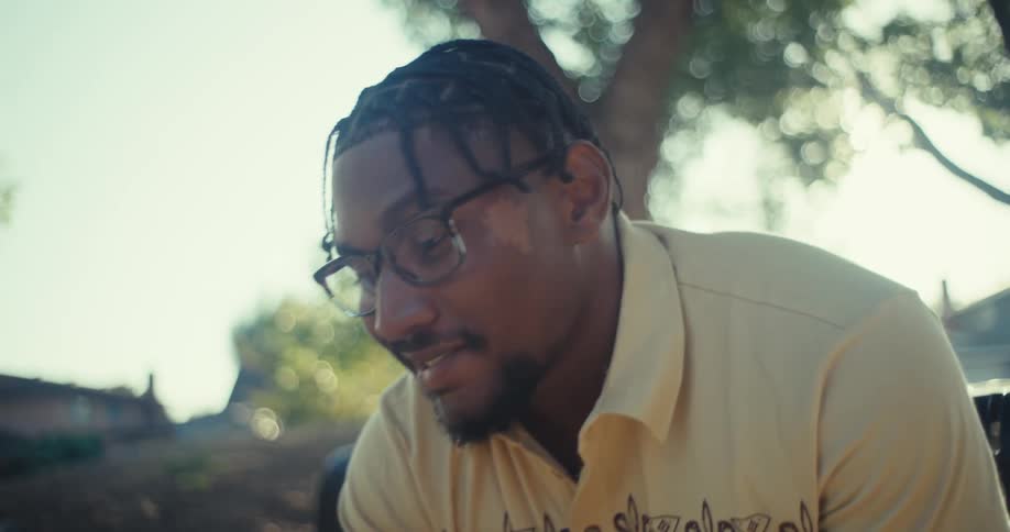 A man with glasses and braided hair sitting on a park bench and engrossed in his smartphone he looks up and smiles suggesting a positive interaction or discovery on his phone