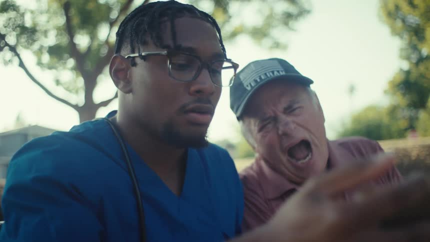 A younger man in blue medical scrubs and glasses sits on a park bench next to an older man wearing a veteran cap both looking at a smartphone held by the younger man they react with surprise and excitement to what they see on the phone