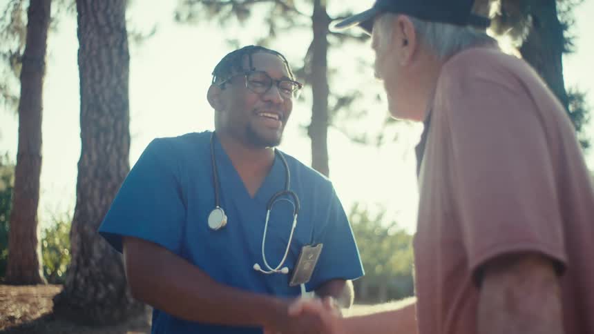 A nurse and an elderly man share a warm handshake and conversation outdoors