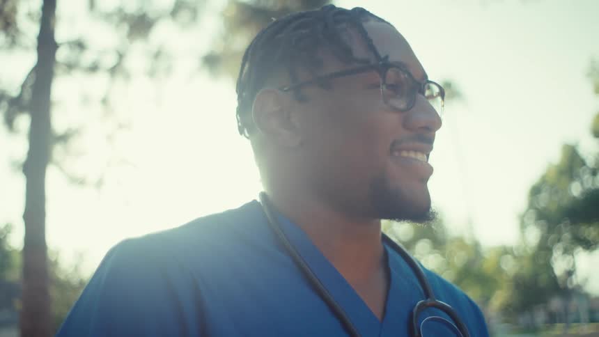 A person wearing blue medical scrubs and a stethoscope stands outdoors with trees in the background