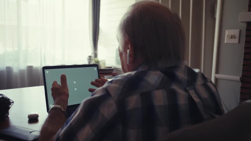 Close up of an elderly man on a call while seated in front of a laptop at a table in a living room during daytime