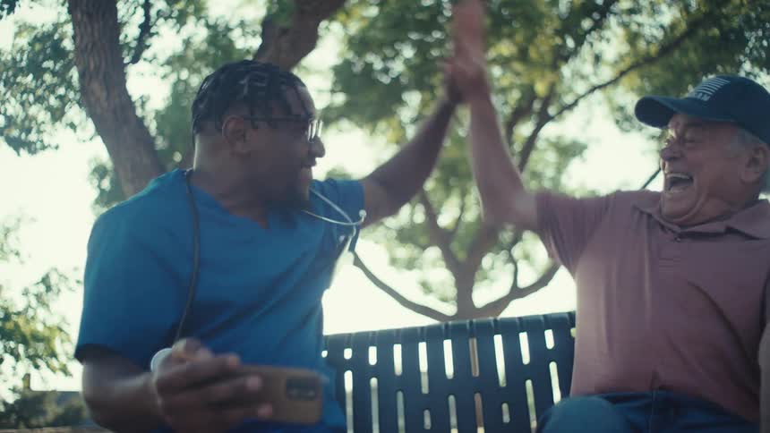 A young caregiver and an elderly man share a joyful high five over a phone screen outdoors