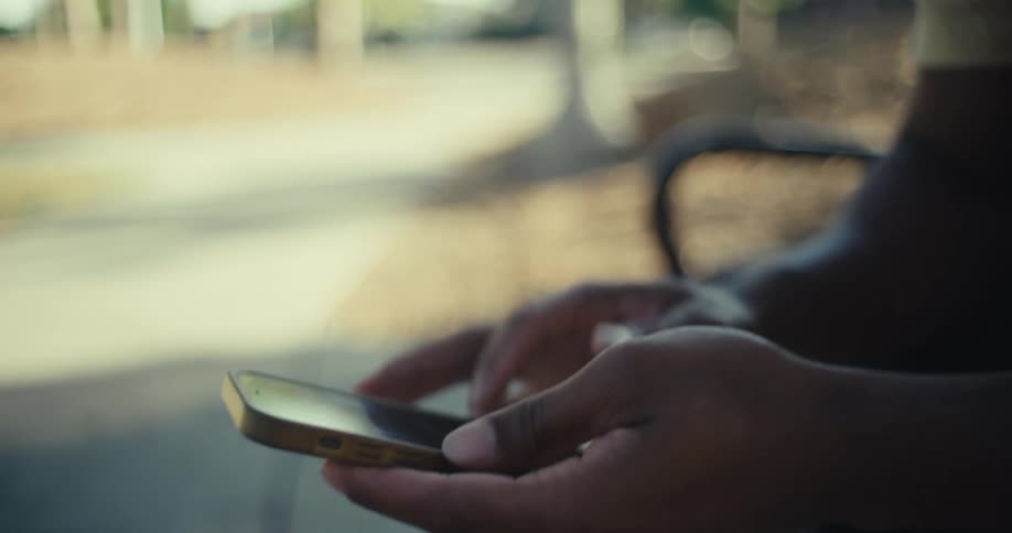 A man is sitting on a bench outdoors scrolling through his phone at midday