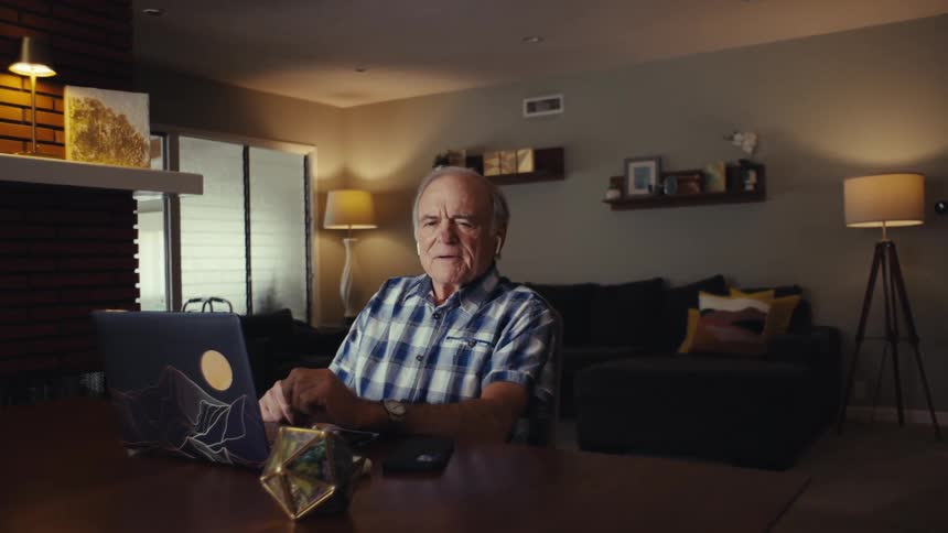 An elderly man in a plaid shirt laughs while seated at a table on a video call wearing earbuds and facing a laptop