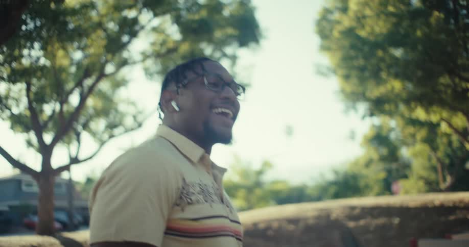 A man with braided hair glasses and white wireless earbuds is seen outdoors holding a smartphone he smiles looking around and occasionally looks up towards the bright sky