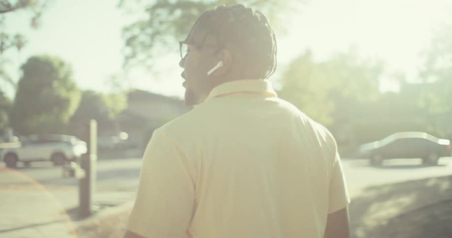 A man with braided hair wearing glasses and white wireless earbuds walks outdoors on a sunny day he is seen from various angles including closeups of his face and shots from behind as he moves along a path with trees and some distant buildings