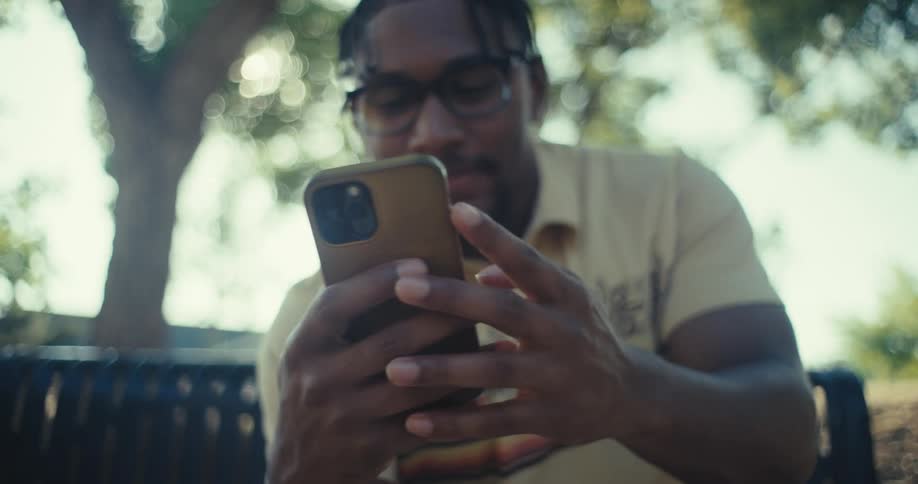 A man smiles while looking at his phone in a sunlit outdoor setting