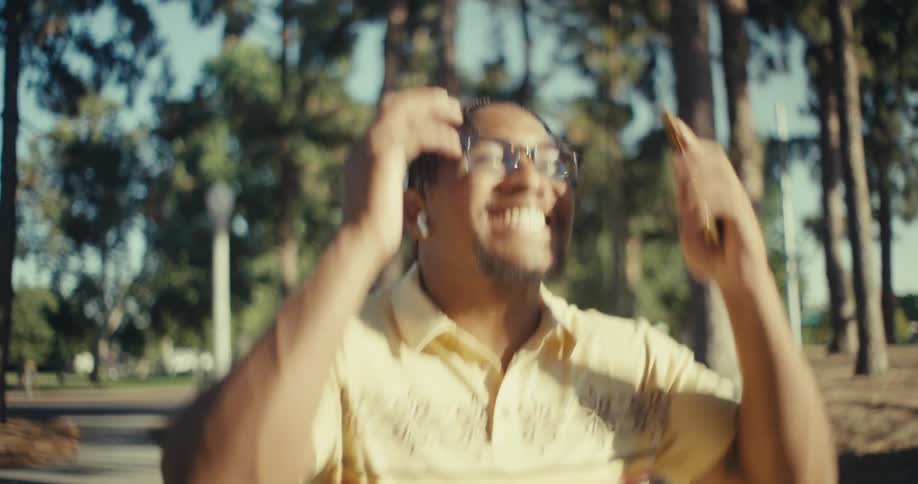 Young black man reacts happily while making a phone call as he walks through a park outdoors during daytime