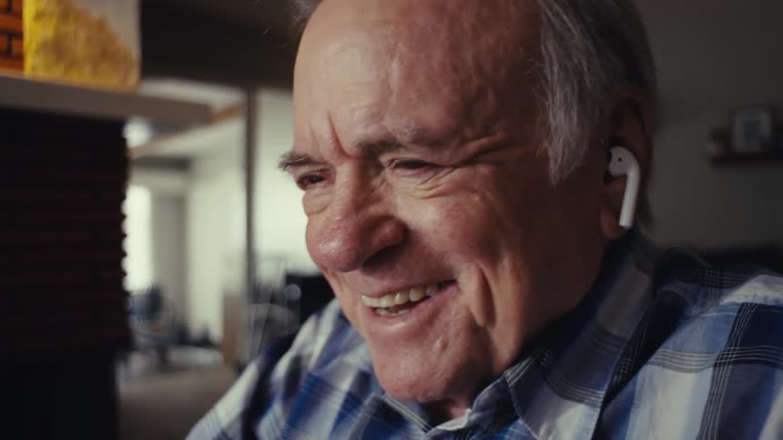 An elderly man wearing wireless earbuds appears to be engaged in a video call in a warmly lit room