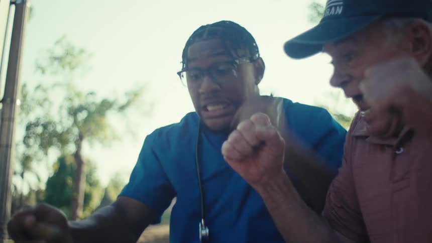 A healthcare professional in blue scrubs with a stethoscope sits beside an elderly man on a wooden bench both erupting with joy as they look at a phone