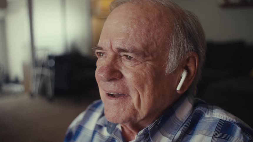 An elderly person in a plaid shirt and white earbuds sits indoors engaged in a phone call