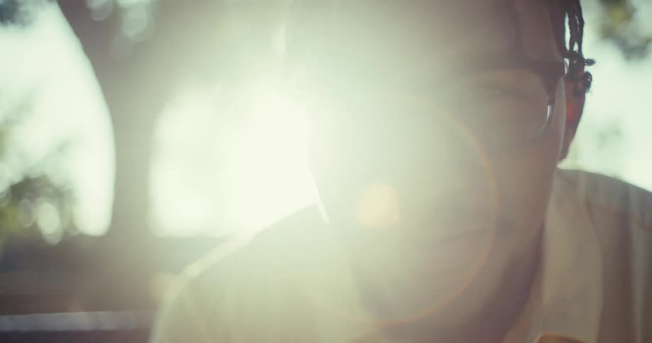 In a bright sundrenched close up a young black man with glasses and braided hair smiles warmly and looks down