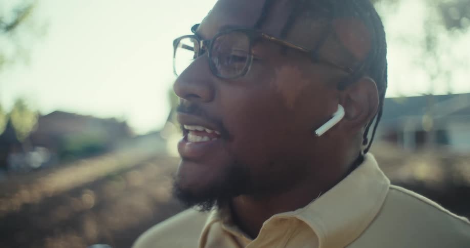 A man wearing glasses and a colorful polo shirt sits on a park bench outdoors smiling while talking on his smartphone with trees and sunlight visible in the background