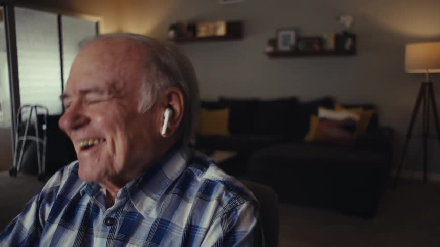 An elderly man wearing earbuds is sitting in his living room laughing