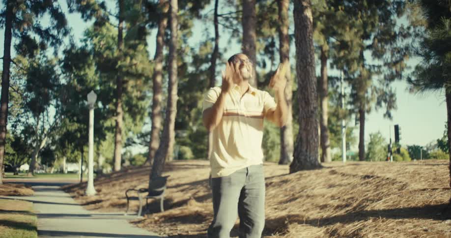 A man with braided hair glasses and white wireless earbuds walks outdoors on a sunny day he is holding a smartphone and appears to be dancing or moving rhythmically smiling and looking at the phone